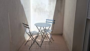 A small outdoor patio with a table and chairs, a window with plants, and a tiled floor.