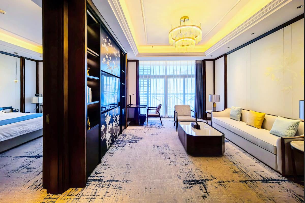 Habitación de hotel con cama, sofá, mesa de centro, silla, estantería y un ventanal grande.