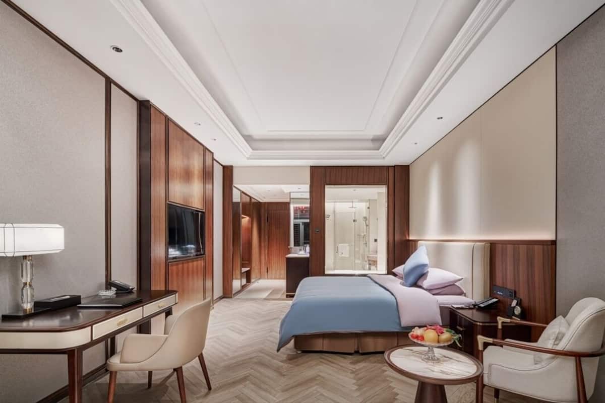 Habitación de hotel con cama, escritorio, silla, televisor y baño visible a través de una puerta abierta.