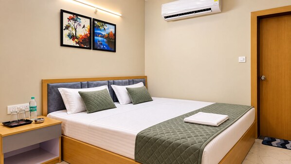 A neatly made bed with a quilted green blanket and two pillows, a bedside table with a bottle and glasses, and two framed pictures on the wall. - The Valoria, Vijay Nagar, Indore (Indore)