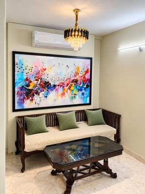 A room with a sofa, a glass coffee table, a wall-mounted air conditioner, and a chandelier.