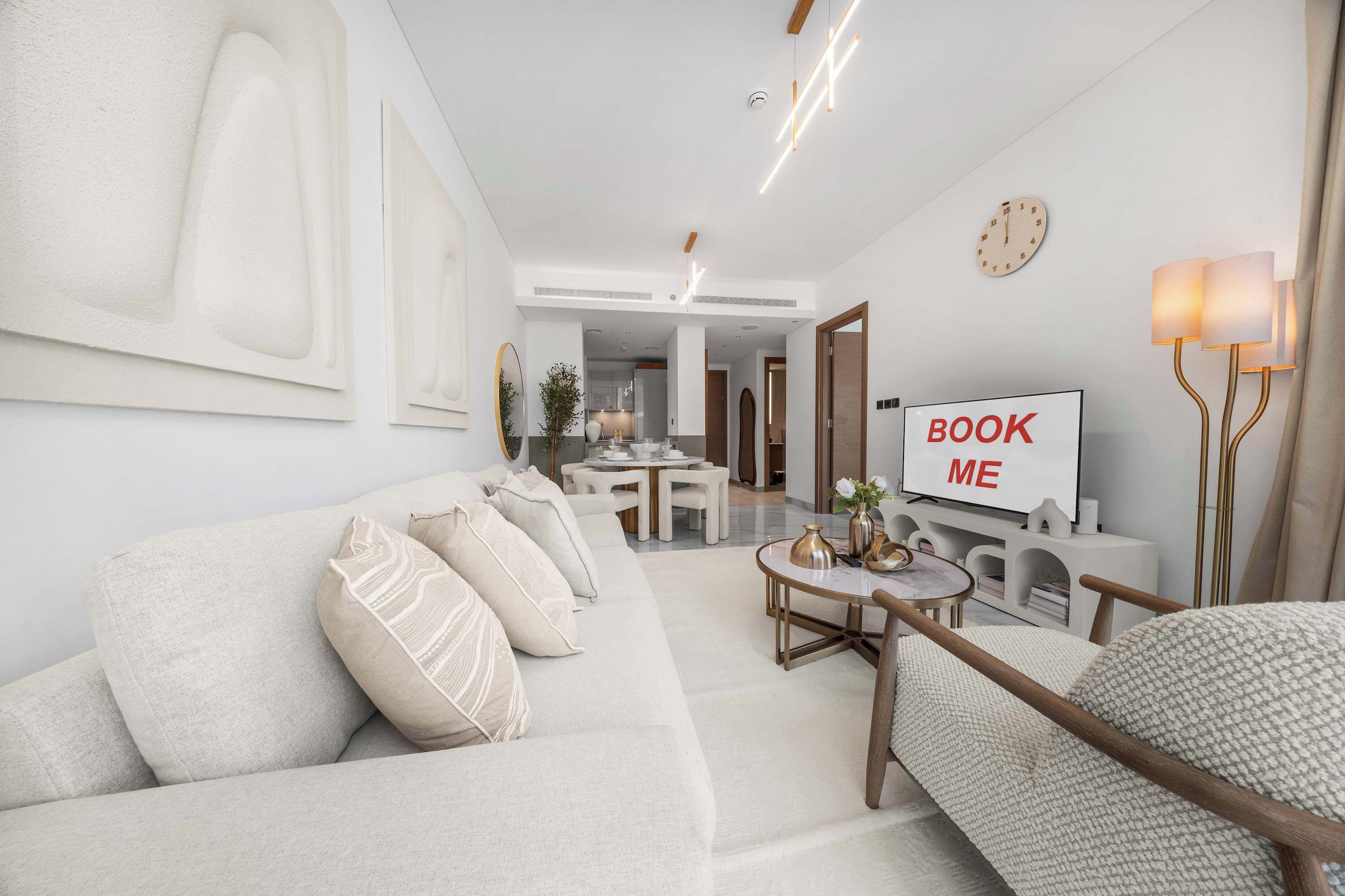 A modern living room with a sofa, a coffee table, a TV displaying 'BOOK ME,' and a dining area in the background.