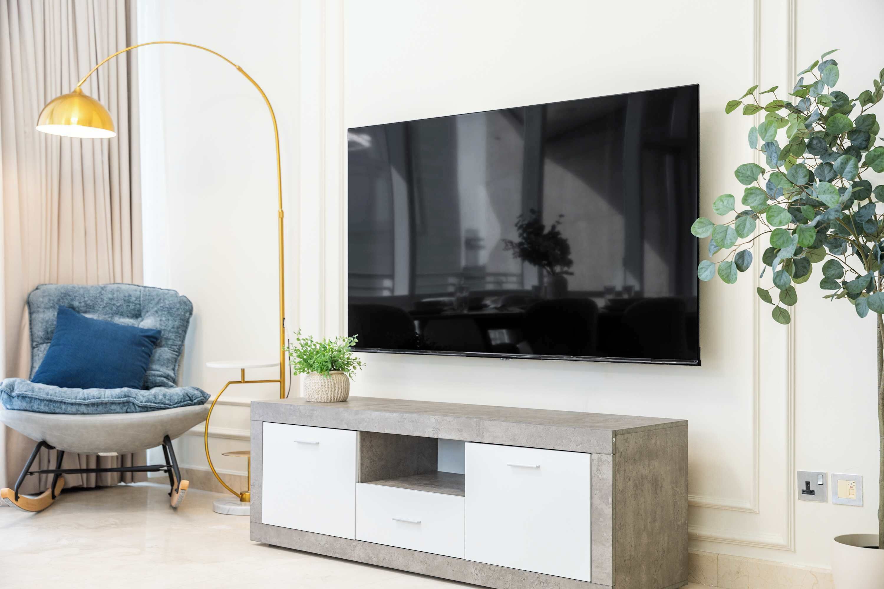 A modern living room with a mounted flat-screen TV, a grey sofa with a blue cushion, a floor lamp, a console table with drawers, and a potted plant.