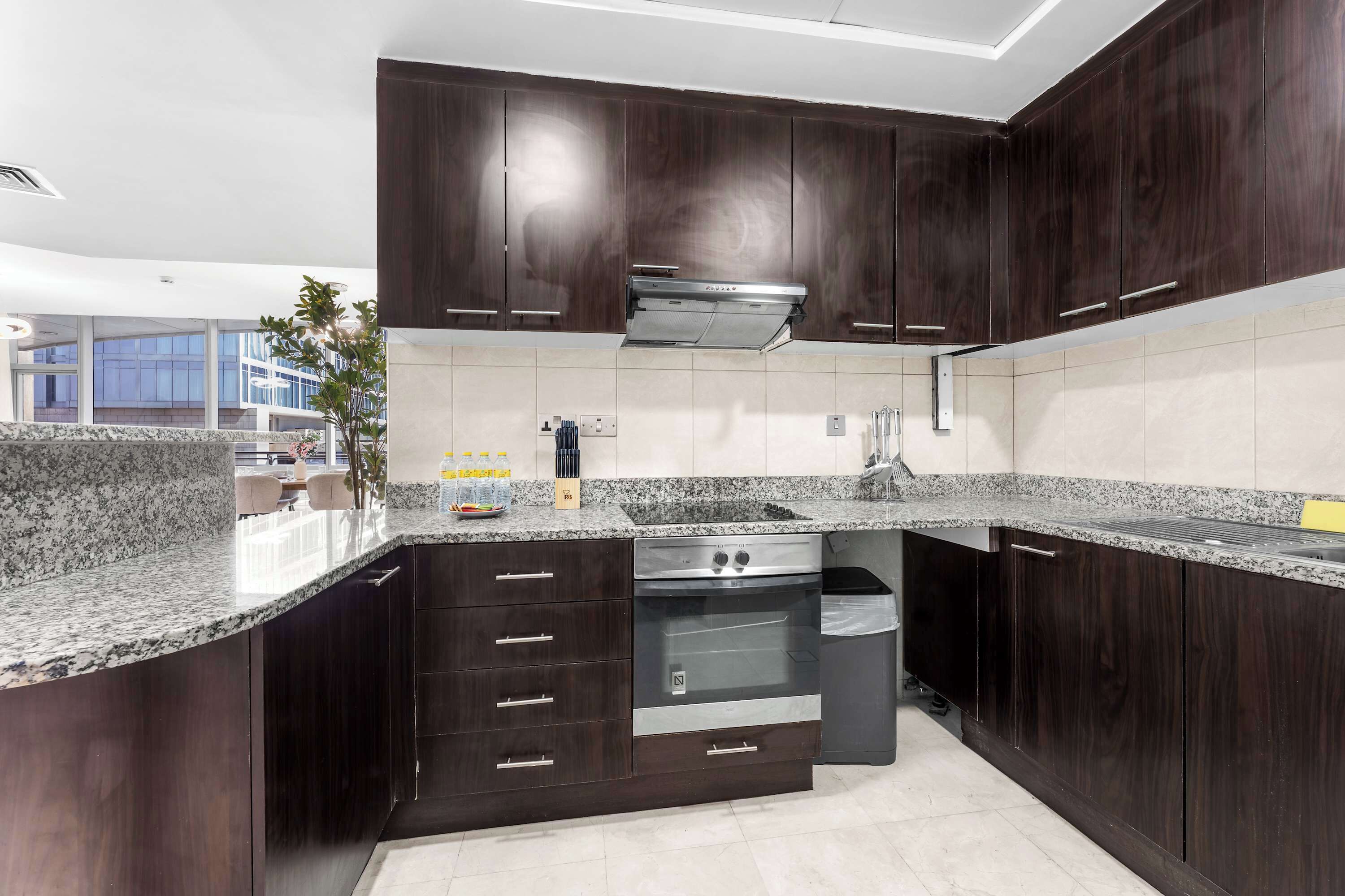 A modern kitchen with dark wood cabinets, granite countertops, a stainless steel range hood, and a built-in oven.