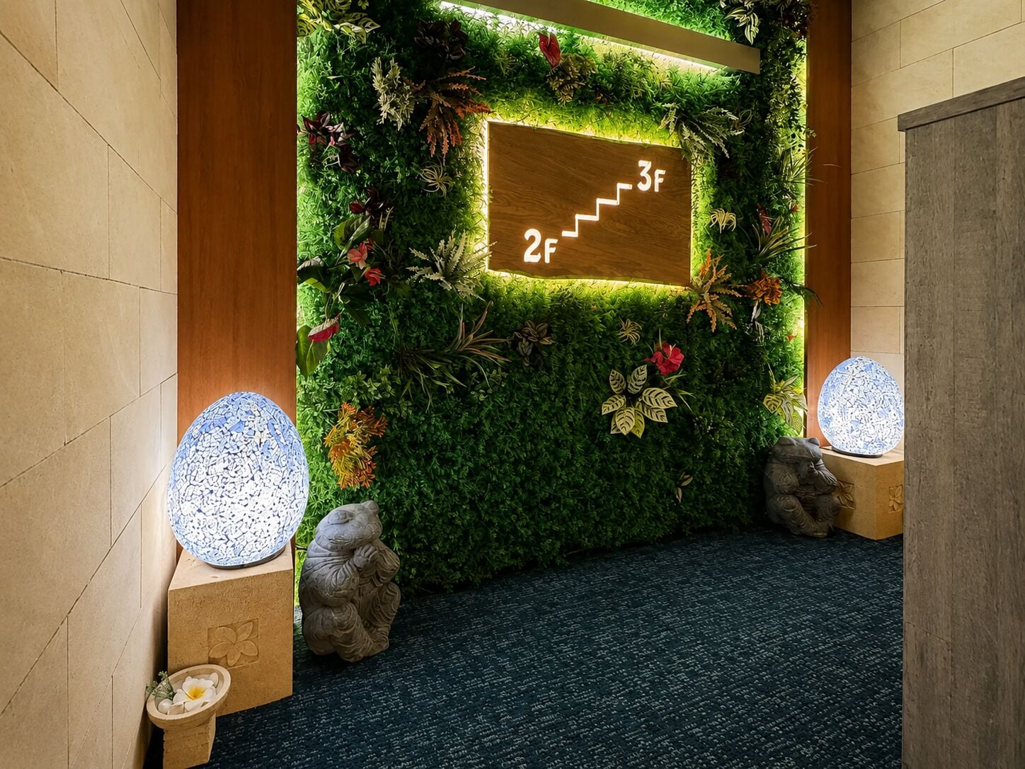 A hallway with a green wall decorated with plants and flowers, a wooden sign with steps, and two stone sculptures on pedestals.