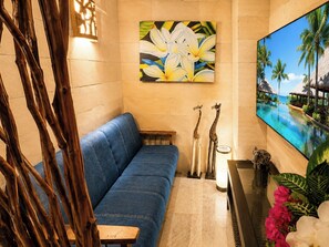 A room with a blue sofa, a painting of flowers, a TV screen with a beach scene, and a small table with a lamp and decorative items.