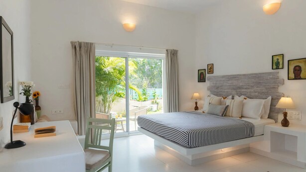 A bedroom with a bed, bedside tables, a desk, a chair, and a view of the outdoors. - Aroha Palms Luxury Apartments (Mandrem)