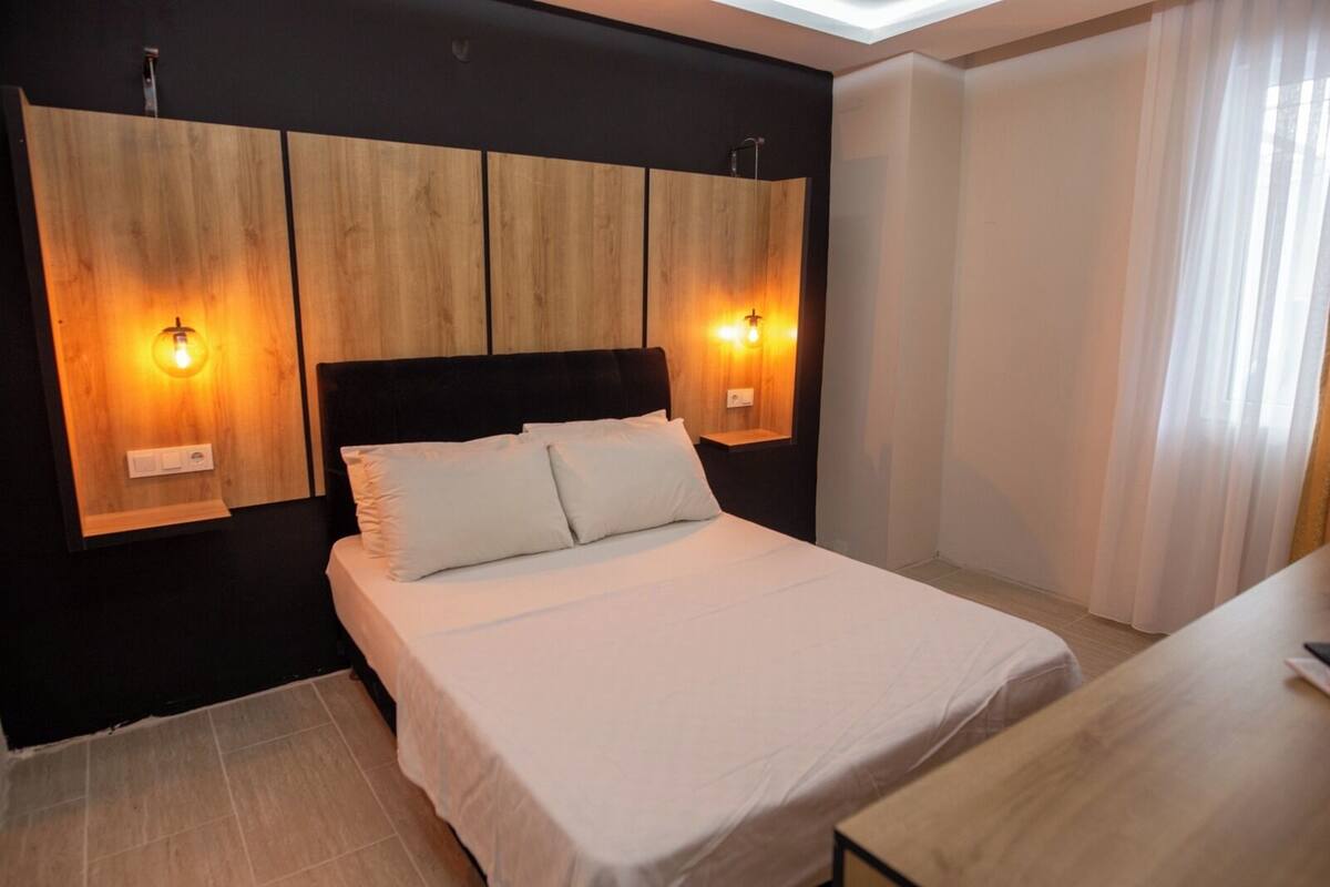 A bedroom with a bed, wooden headboard, two wall-mounted lights, and a window with curtains.