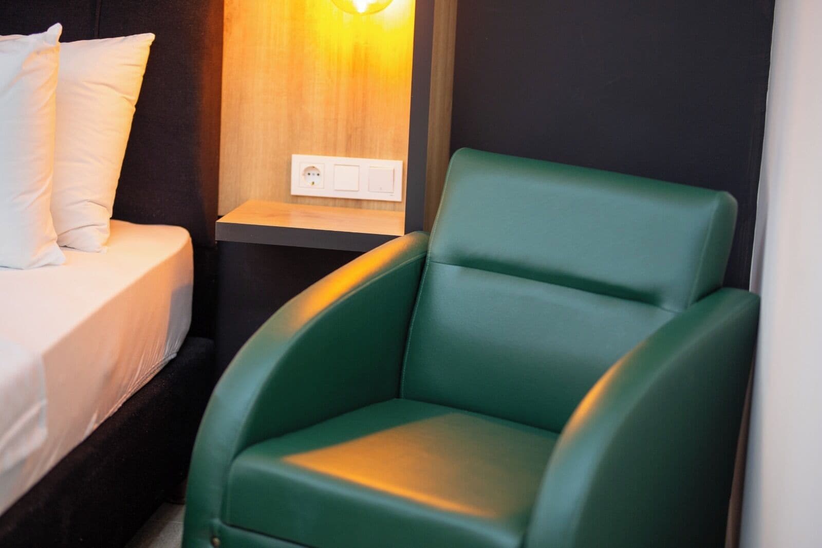 Una habitación con una cama, una silla verde, una mesita de noche con luz y un enchufe.