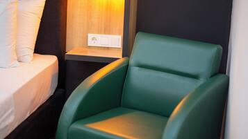 Una habitación con una cama, una silla verde, una mesita de noche con luz y un enchufe.