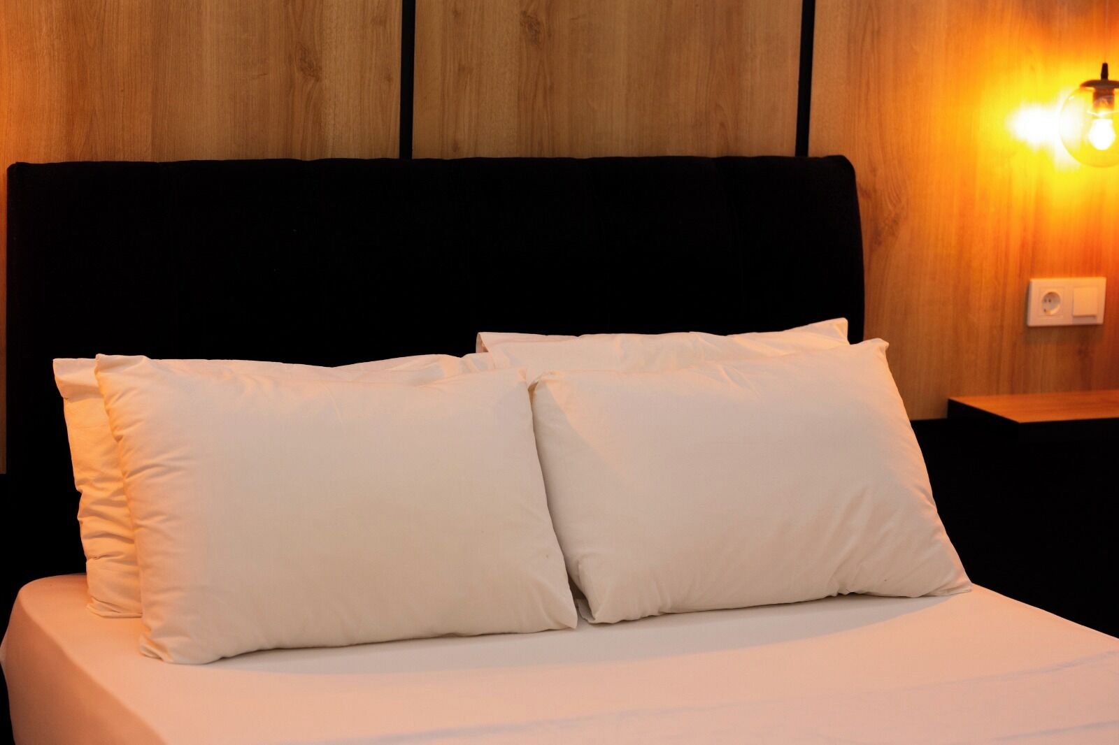 Una cama con ropa blanca y almohadas, cabecera de madera y lámpara fijada a la pared.