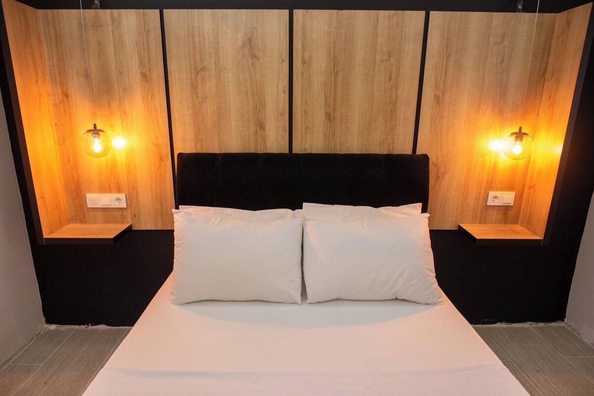 A bed with two white pillows, a black headboard, and wooden panel walls with two wall-mounted light fixtures.