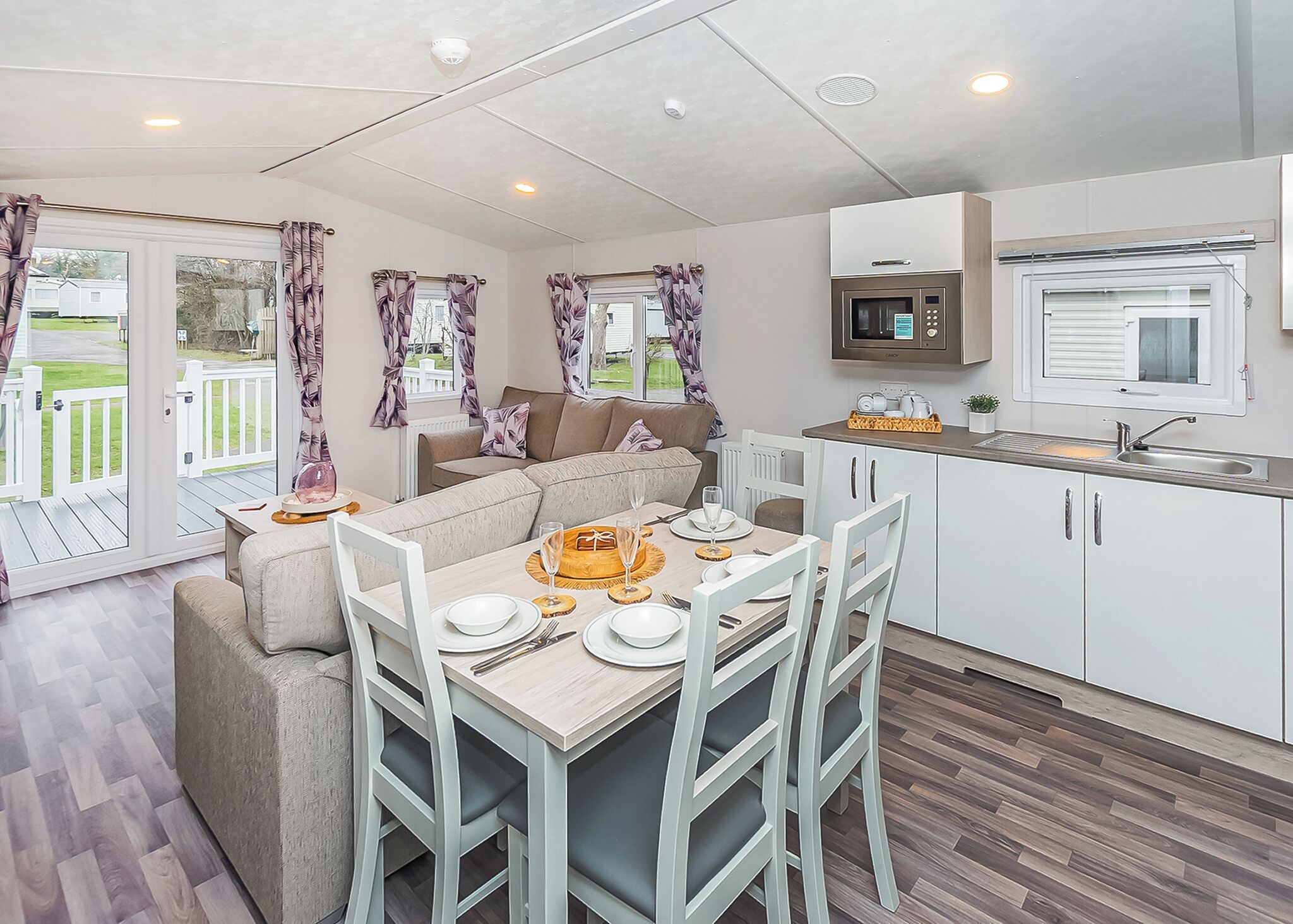 A modern kitchen with white cabinets, a microwave, and a sink. There is a dining area with a table set for two, and a sofa in the background.