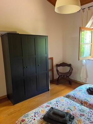 A room with a large wardrobe, a wooden chair, a bed with floral bedding, and a window with a view of trees.