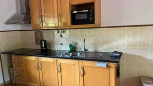 A kitchen with wooden cabinets, a black countertop, a stainless steel sink, a microwave, and a trash bin.