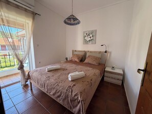 Ein Schlafzimmer mit Bett, Nachttisch, Hängeleuchte, einem Fenster mit Vorhängen und Blick auf einen Balkon.