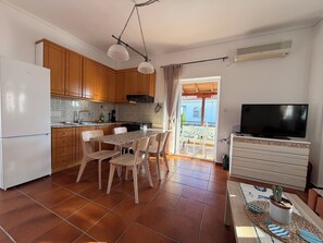Eine kompakte Küche mit Holzschränken, einem weißen Kühlschrank, einem Esstisch mit Stühlen, einem Fernseher auf einem wei ßen Schrank und einer Schiebetür zu einem Außenbereich.