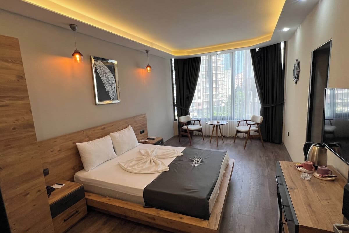 A modern hotel room with a wooden bed, a desk with a kettle, a TV, and a view of the city through large windows.
