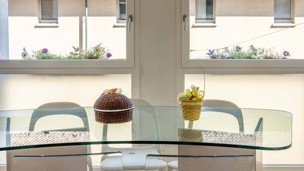 A dining area with a glass table, clear chairs, and a view of buildings through a window. - Lame Pro (Bologna)