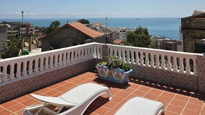 A terrace with two white lounge chairs, a decorative planter, and a view of the sea and buildings.