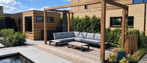 A modern outdoor patio with a pool, wooden pergola, and a grey sofa.