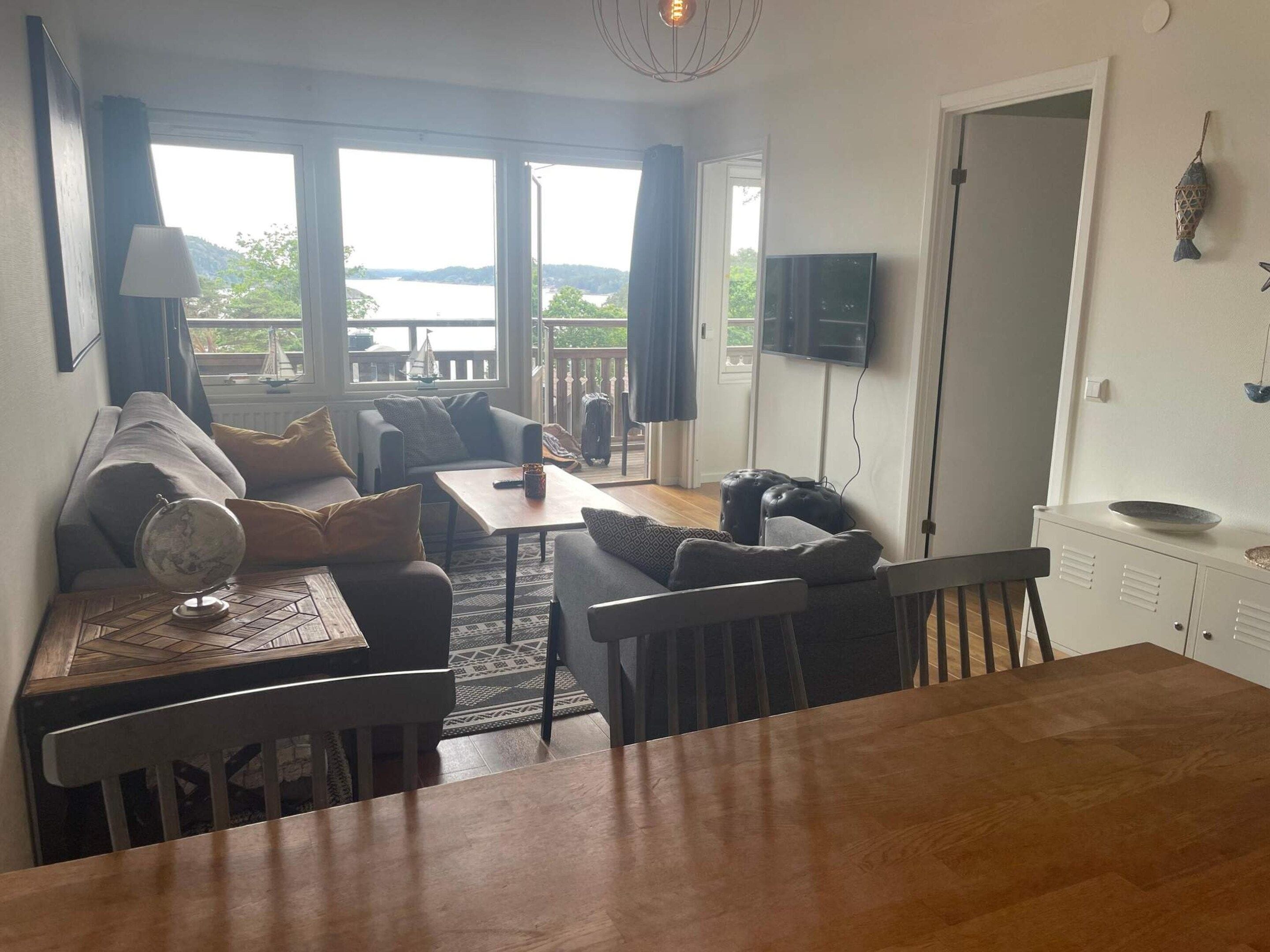 A cozy living area with a sofa, a coffee table, and a dining table. There is a television mounted on the wall and a window with a view of the outdoors.
