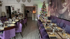 Um restaurante com mesas grandes e partilhadas, cadeiras estofadas em roxo e um pinheiro de Natal.
