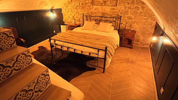 A bedroom with a stone wall, a bed with a headboard, a nightstand, and a wooden staircase.