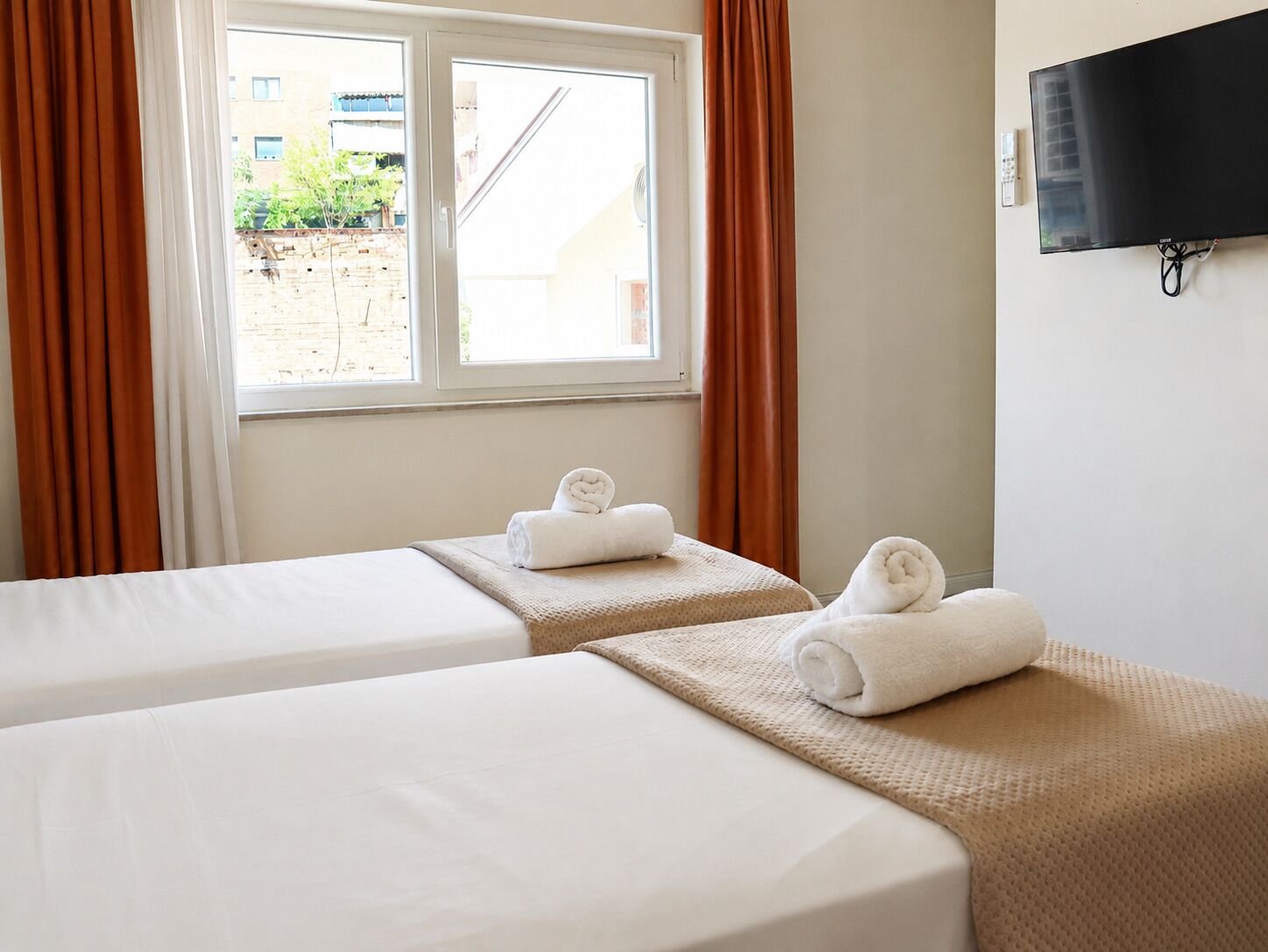 Camera d'albergo con due letti, una finestra con tende e una televisione fissata al muro, con vista sull'esterno.