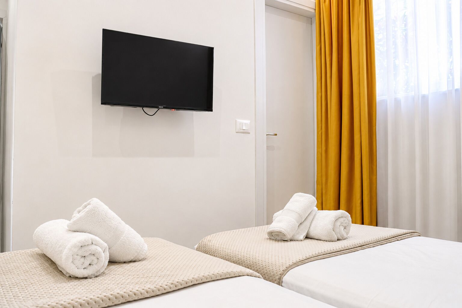 Una camera d'albergo con due letti, una TV a schermo piatto e una finestra con tende gialle.