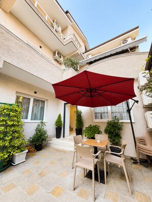 A patio with a table and chairs, a red umbrella, potted plants, and a building with balconies. - Dama Residence (Tirana)