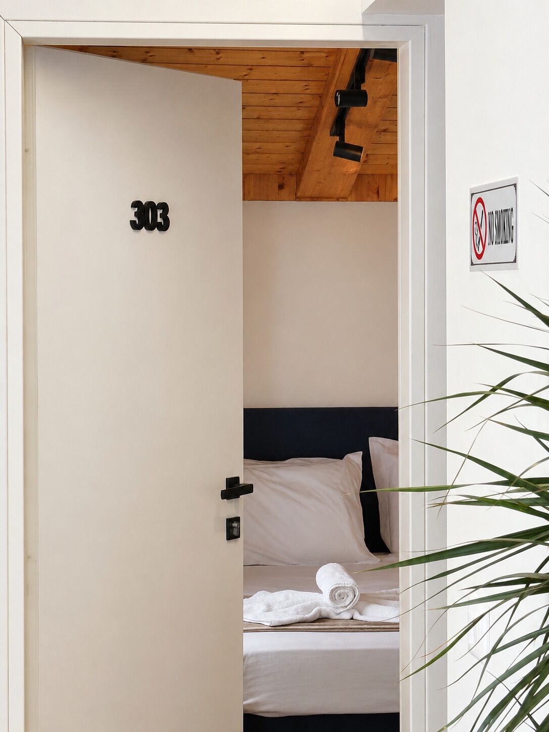 Habitación de hotel con el número 303 en la puerta, un cartel de 'No Fumar', una cama con ropa blanca y una planta.