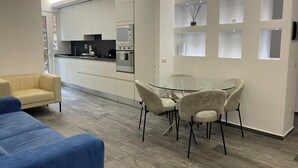 Eine moderne Küche mit weißen Schränken und eingebauten Geräten, ein Glastisch mit vier Stühlen sowie ein gelbes Sofa in einem Wohnbereich.