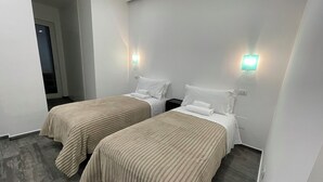 Ein Hotelzimmer mit zwei Betten, jeweils mit einer beigen Bettdecke, weißen Kissen und einem Nachttisch. An beiden Seiten der Betten befinden sich Wandleuchten.