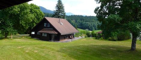 A rustic cabin with a sloped roof, surrounded by a grassy field and trees, with mountains in the background.