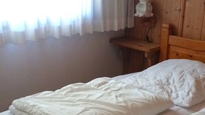 Un lit en bois avec une literie blanche, dans une pièce aux murs en bois et dotée d’une fenêtre ornée de rideaux blancs.