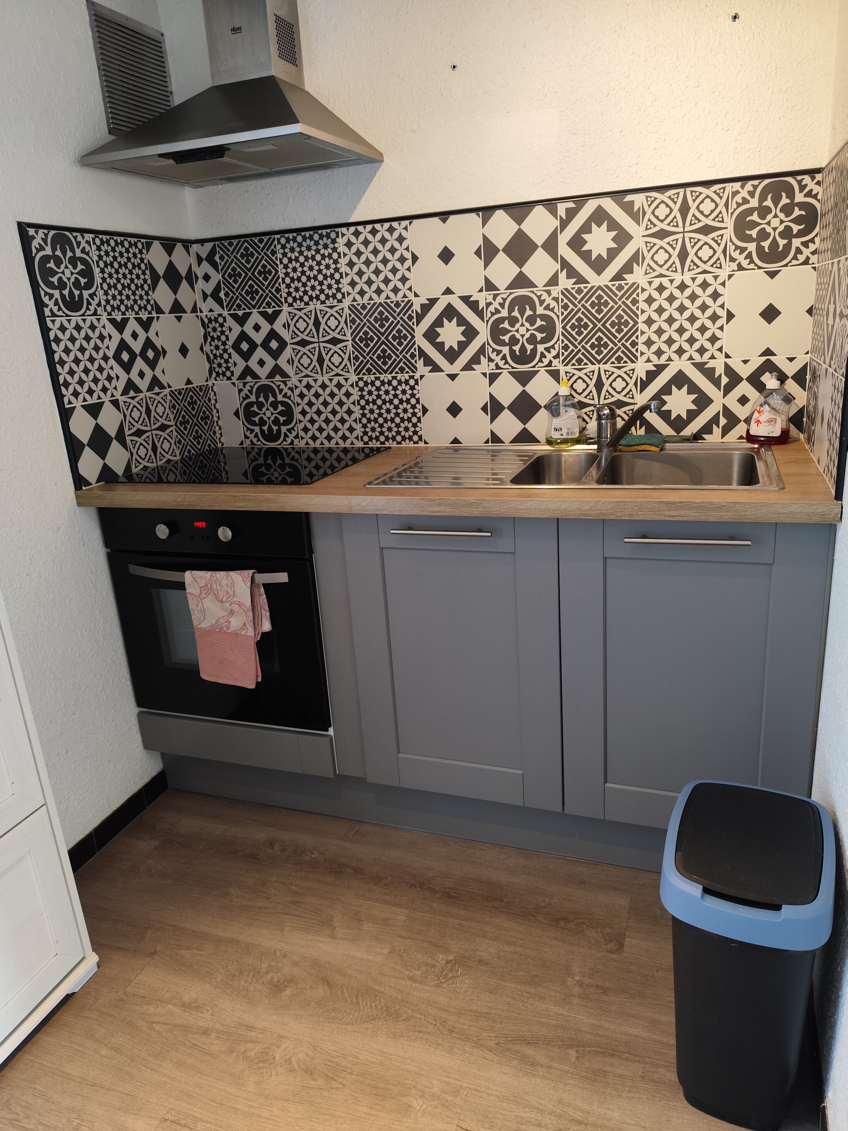 Een keuken met patroontegels, een roestvrijstalen spoelbak, grijze kastjes, een houten aanrechtblad, een zwarte prullenbak en een roestvrijstalen afzuigkap.