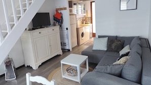 Una sala de estar con un sofá gris, un mueble de televisión blanco, una cocina al fondo y una escalera.