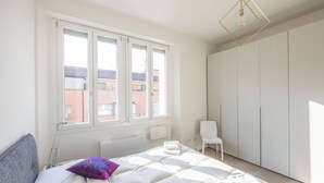 Een slaapkamer met een bed, een stoel, een kledingkast en een raam met uitzicht op gebouwen.