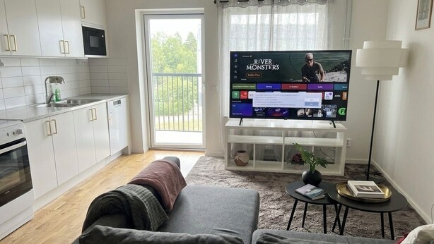 Uma sala de estar moderna com televisão de tela plana, um sofá, uma mesa de centro e uma área de cozinha com armários e eletrodomésticos brancos.