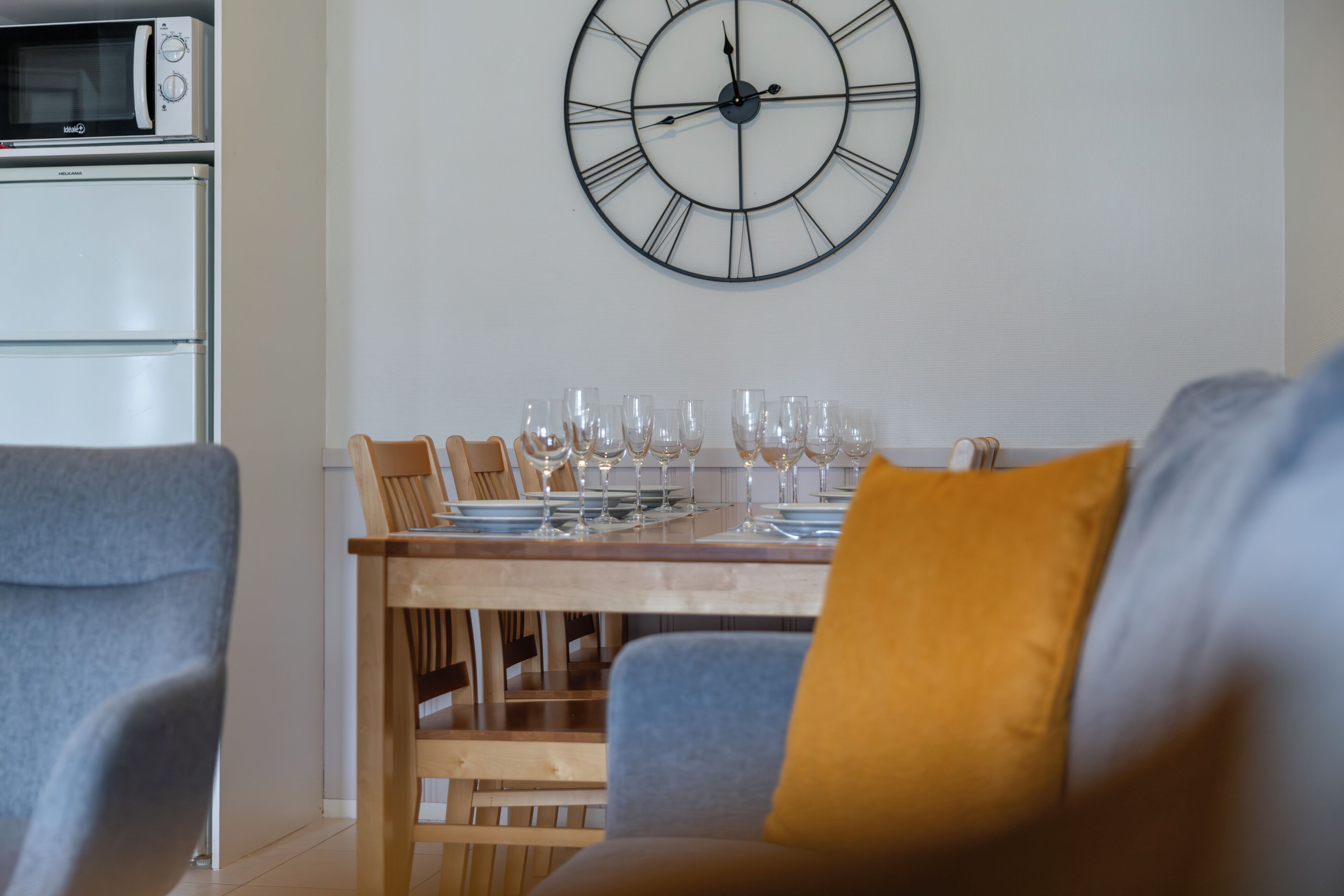 A dining area with a wooden table set for a meal, a large wall clock, and a microwave above the table.