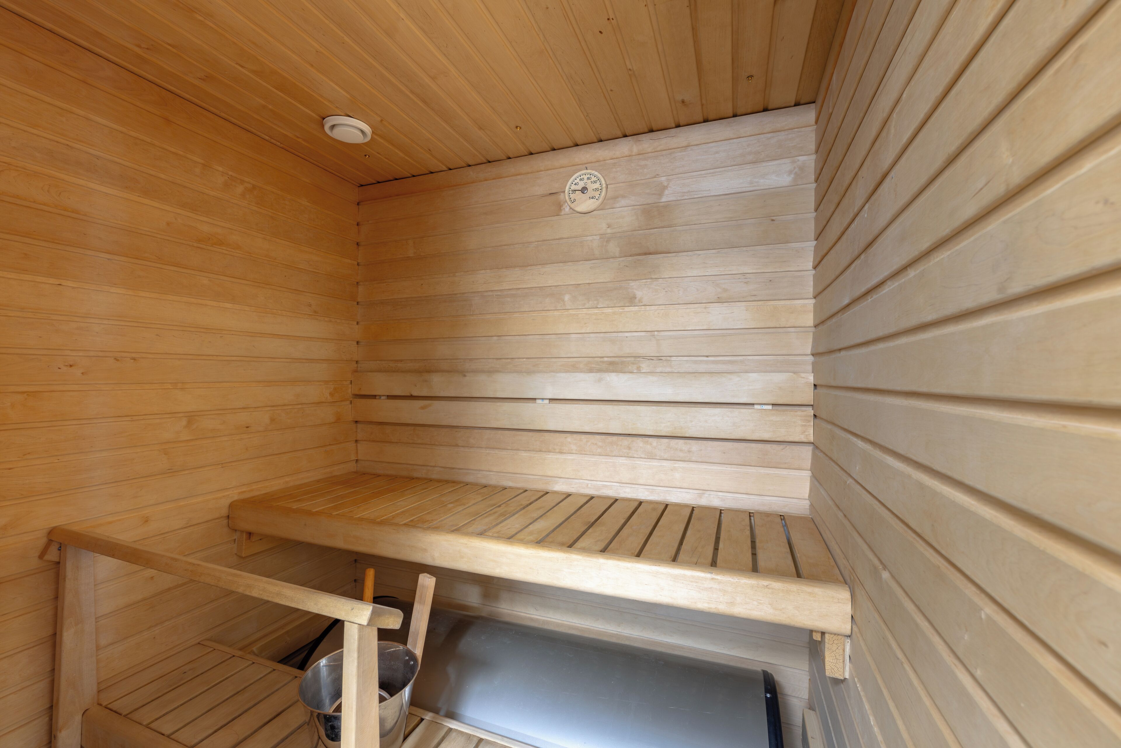 A sauna with wooden walls and benches, a ceiling vent, and a metal bucket.