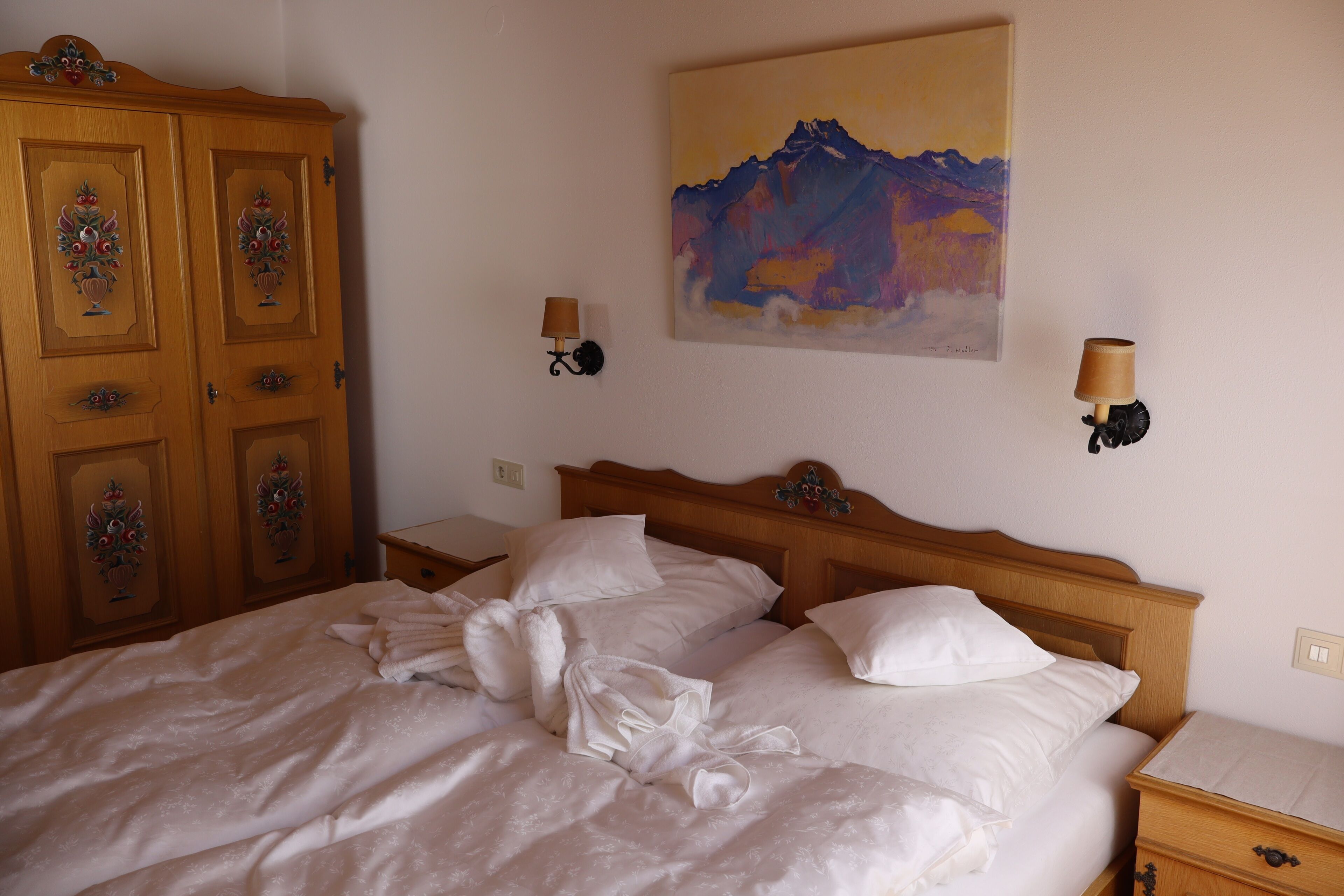 Ein ordentlich bezogenes Bett mit weißen Leinen, ein hölzerner Kopfseitenverkleidung, ein Nachttisch mit Lampe und ein Bergpanorama an der Wand.