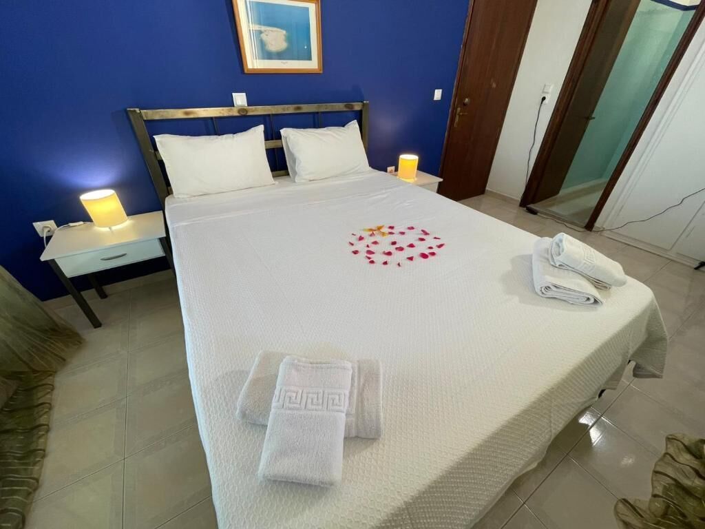 Un lit bien fait, recouvert d’une couvre-lit blanche, avec deux oreillers et un motif décoratif en forme de cœur. On y trouve deux lampes de chevet, une serviette pliée et un téléphone.