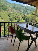 Terraza de madera con una mesa y dos sillas, con vista a un paisaje montañoso con casas.