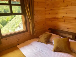 A wooden cabin room with a bed, two pillows, a window with curtains, and a view of greenery outside.