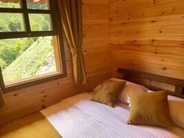 Habitación de cabaña de madera con una cama, dos almohadas, una ventana con cortinas y vistas al verde exterior.