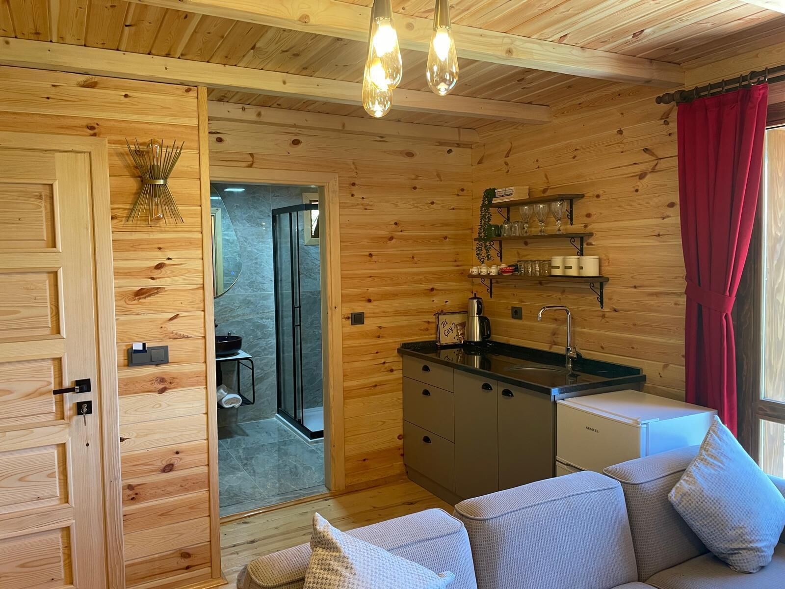 Interior de una cabaña de madera con kitchenette, sofá y zona de ducha.