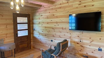 A wooden-paneled room with a flat-screen TV mounted on the wall, a cushioned chair, a small table, and a red bedspread.