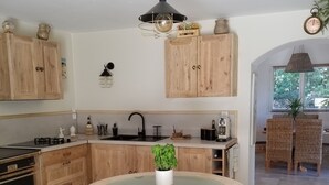 A kitchen with wooden cabinets, a round glass table, wicker chairs, and a hanging light fixture. - A haven of peace (Néoules)